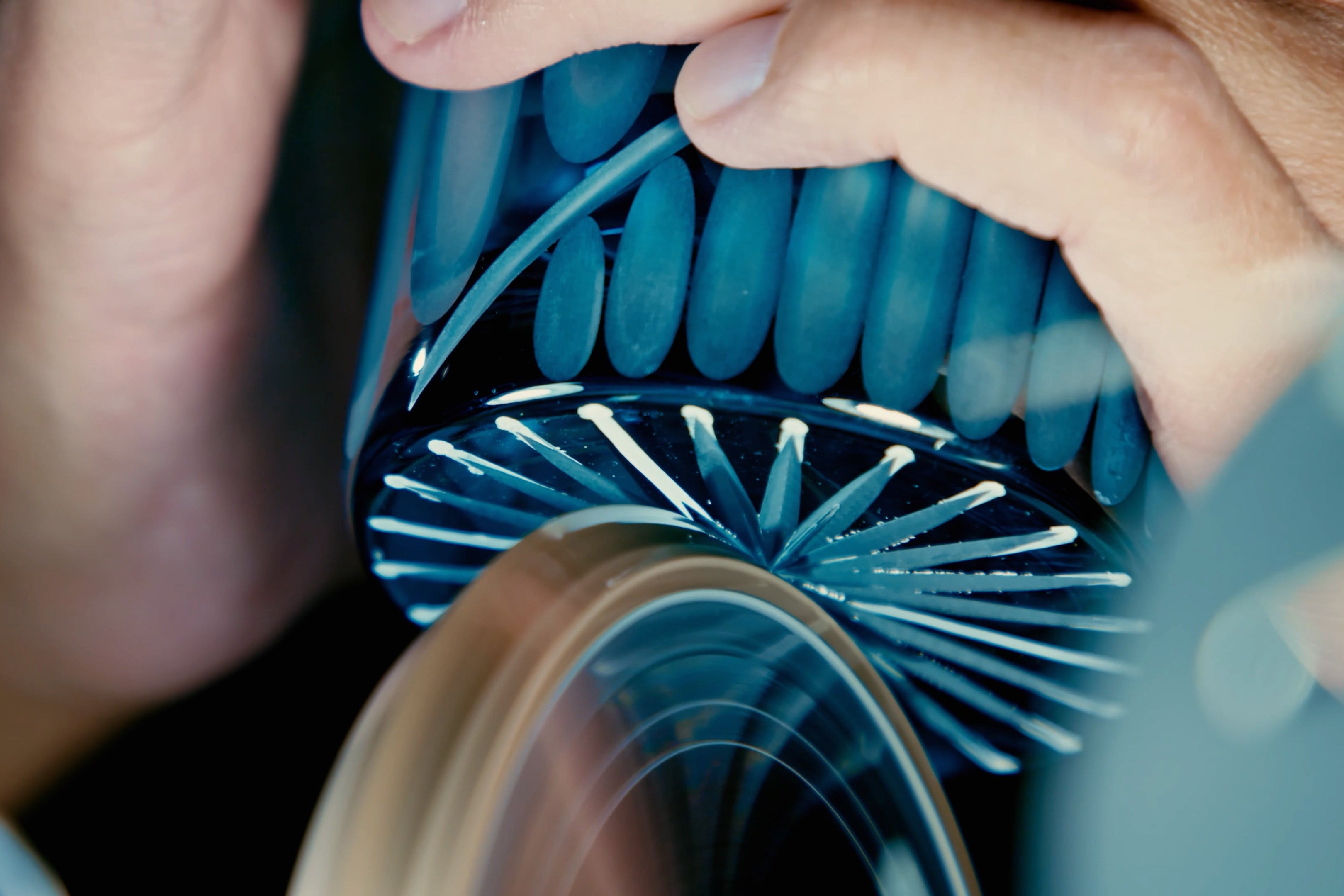 An artisan is making cut patterns on a blue glassware against a wheel